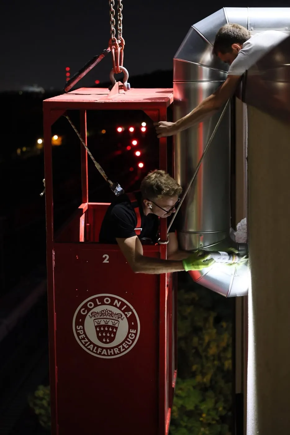 Monteur im Krankorb neben einer angehobenen Wärmepumpe bei Nacht