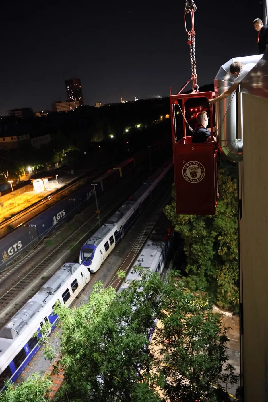 Wärmepumpe am Kranhaken über den Gleisen, im Hintergrund Köln bei Nacht