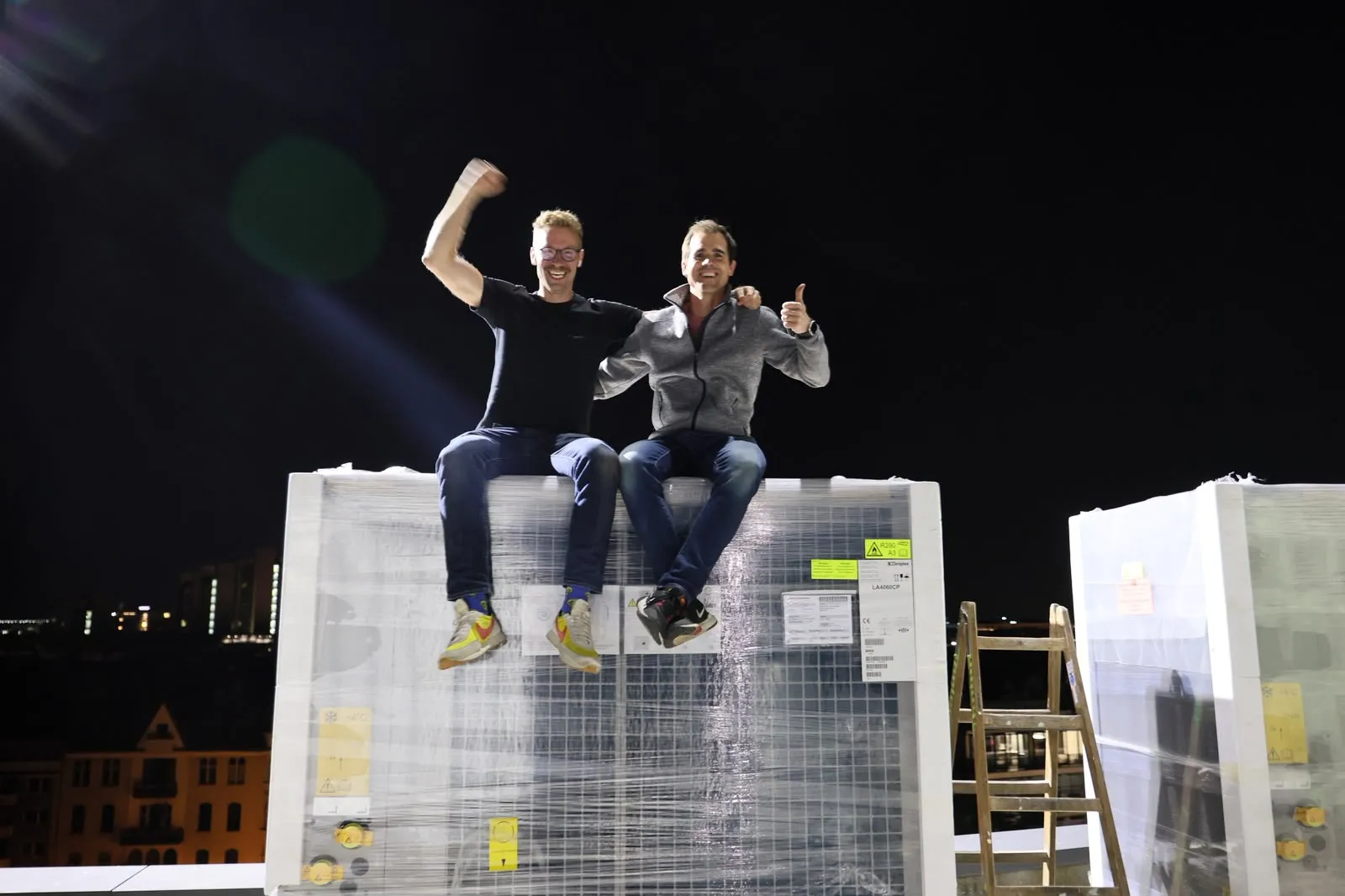 Tilman Gartmeier und Patrick Mexner sitzen nachts auf einer noch eingeschweißten Wärmepumpe auf dem Dach des Stadtgarten-Karrees, im Hintergrund die Lichter von Köln