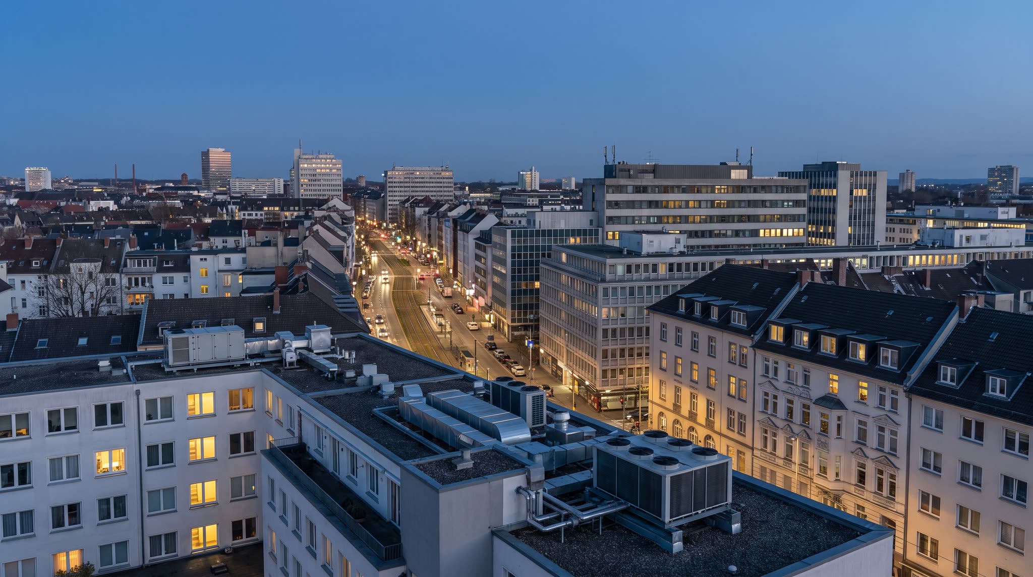 Blick über ein deutsches Stadtviertel in der blauen Stunde, im Vordergrund ein Flachdach mit Lüftungs- und Wärmepumpenanlagen