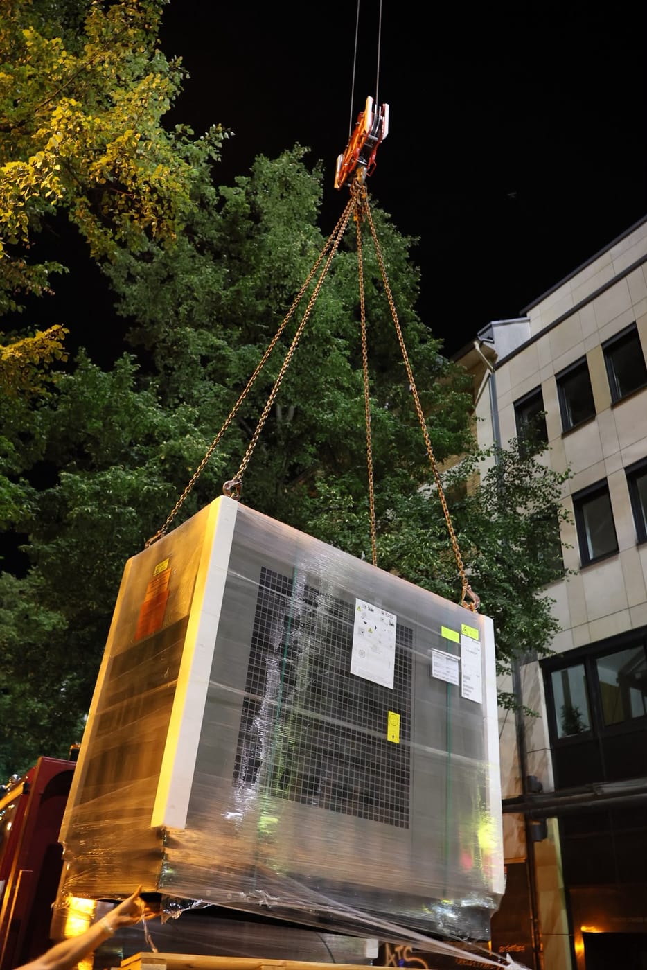 Eine Wärmepumpe hängt nachts am Kran über der Venloer Straße in Köln und wird auf das Dach des Stadtgarten Karrees gehoben